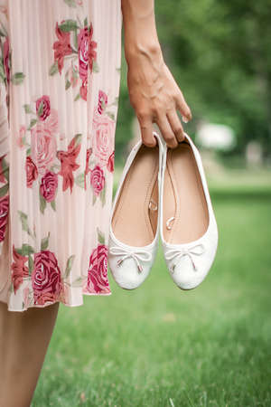 Young pretty woman in summer dress walking on meadow on green grass. A young girl holds white ballet shoes in her hands.の写真素材