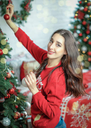 Happy young brunette female decorating christmas tree. Girl holding a christmas ball in her hands and looking into cameraの写真素材