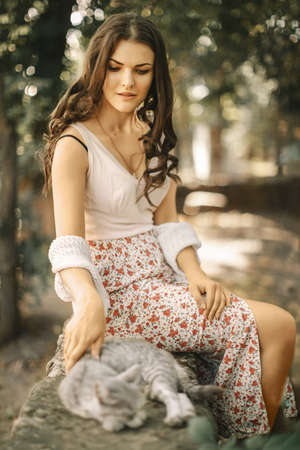 Retro style portrait of pretty young woman caressing a little gray kitten sleeping on a stone fenceの写真素材