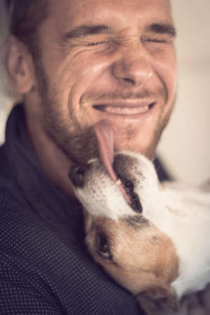 Close-up portrait of young attractive smiling guy and purebred dog jack russell terrier licking mans...