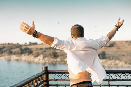 A portrait from the back of a man with his arms raised up against a background of sky and water. The man is wearing a white open shirtの写真素材