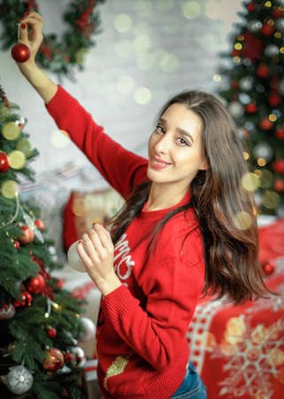 Portrait of happy young brunette female decorating christmas tree. Girl holding a christmas ball in her hands and looking into cameraの写真素材
