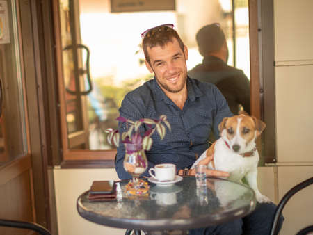 Portrait of young smiling man drinking coffee in cafe together with cute purebred dog jack russell terrier sitting on his kneesの写真素材