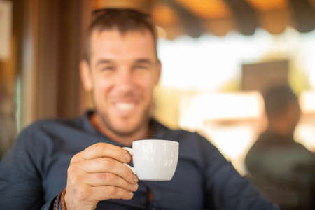 Blurred portrait of young smiling man sitting in cafe and drinking coffee. Focus on white cup in his hand.の写真素材
