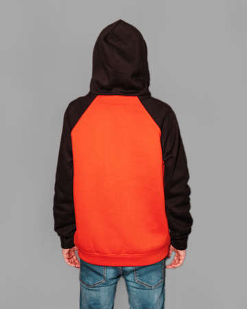 A young guy dressed in a orange and black hoodie with a hood on his head standing backwards.の写真素材