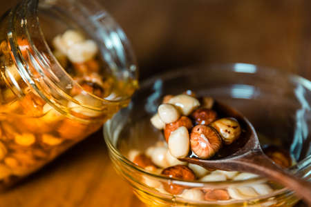 Farmer Honey mixed with nuts, seeds pouring from a transparent jar into a wooden spoon on glass plate on kitchen wooden table. Still life closeup photographyの写真素材