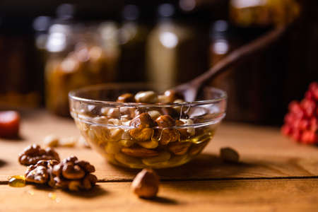 Honey mixed with nuts, seeds in a transparent plate with wooden spoon, one jar, walnuts kernel, red rowan, dried apricots, piece of pumpkin on rustic wooden kitchen table. Still life photography.の写真素材