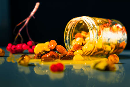 Closeup jar of honey mixed with nuts and rowan berries spilled on a reflective surface on black...