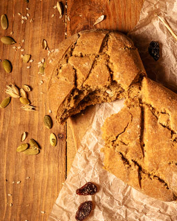Close-up broken homemade fresh bread, pumpkin seeds and raisins on parchment paper wooden table.