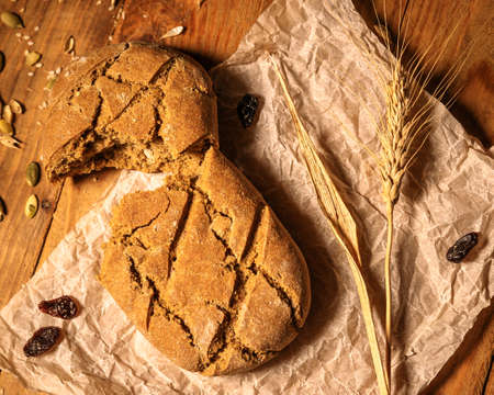 Close-up broken homemade fresh bread, ear of ripe wheat, pumpkin seeds and raisins on parchment paper wooden table.の写真素材