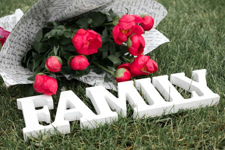 Wooden white word Family and bouquet of pink peony flowers on green grass background outdoors, copy...