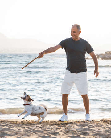 60-years old man playing with small cute jumping and barking dog jack russell terrier on sea beachの写真素材