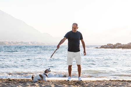 Elderly 60-years old man playing with small cute dog jack russell terrier on beach. Person and animal friendshipの写真素材