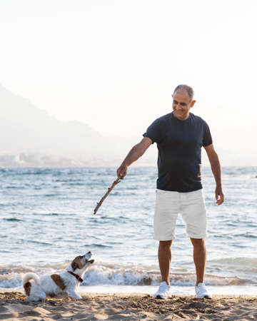 Portrait of elderly 60-years old man playing with small cute dog jack russell terrier on beach.の写真素材