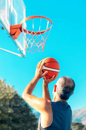 60-years old basketball player shoots hoops outdoors. Elderly amateur man throws a leather ball into...