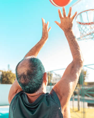 Aged basketball player shoot hoops outdoors. Elderly amateur man throws a leather ball into a basket on a sunny summer day.の写真素材