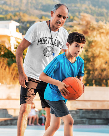 Northen Cyprus, Girne, Alsancak - Jule, 25, 2023: Basketball sport training for children teenagers around 7-14 years old with an elderly coach on a basketball court in summer.のeditorial素材