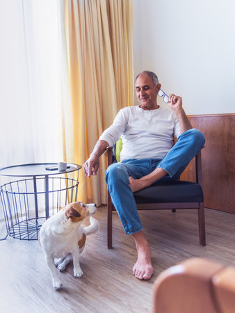 Smiling senior man sits in room and plays with terrier with ballの写真素材