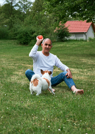 Senior Man Sitting on Grass Teasing Dog with Ballの写真素材