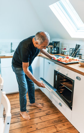 Senior Man Opening Oven Door Cooking Vegetarian Meal in Modern White Kitchen with Copy Spaceの写真素材