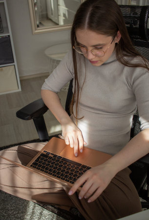 Young woman sitting in office chair working on laptop, focused typing posture, modern home interior...