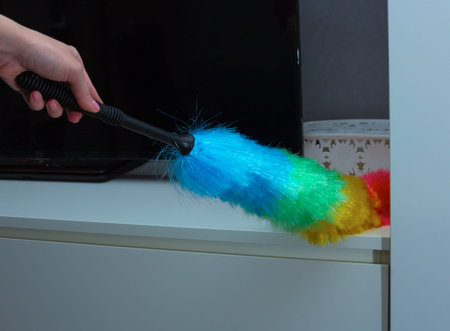 Woman cleaning white cabinet surface with colorful feather duster in modern living room