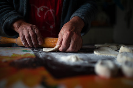 Old female hands at work in the kitchen close up. Grandmother rolls out a piece of dough with a wooden rolling pin. Cooking process. High quality dark photoの写真素材