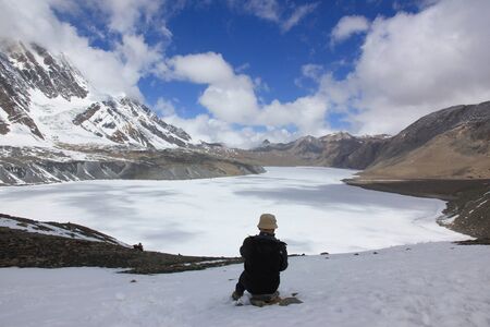 A man sits with his back to the camera by a frozen lake in the mountains of Nepal.の写真素材
