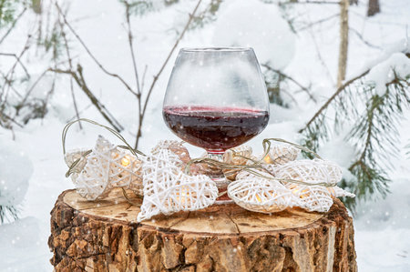A glass of wine and a garland of hearts on a wooden stump in a snowy forestの写真素材