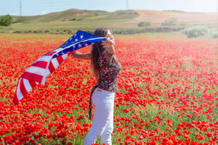 Girl in white clothes with an flag in her hands enjoys the wind and rays of the sun of a amazing red blooming poppy fieldの写真素材