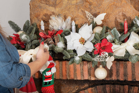 hands decorate festive a Christmas tree garland with lights, glowing bulbs, toys, bows, knitted shoes, balls, poinsettia flowers and candles on a stone fireplace, close-up, selective focusの写真素材