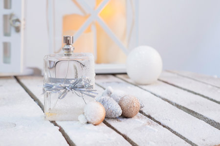 a bottle of perfume tied with a festive ribbon on a snowy wooden surface with balloons with cones against the background of blurry lantern lights and white balloons in the background, selective focus.の写真素材