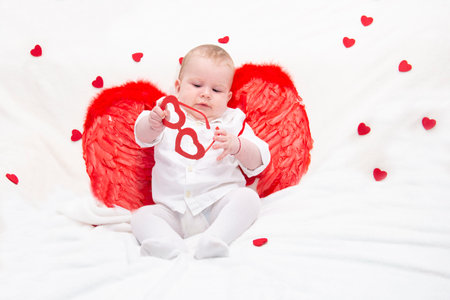 A baby with puffy cheeks in white clothes with red cupid wings, plays with glasses with hearts, isolated on a white tender blanket with hearts. cupid kid for a project with lovers.の写真素材