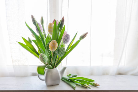 a bouquet of tulips in a ceramic teapot with decorative colored eggs on the window sill. Home decor for Easterの写真素材