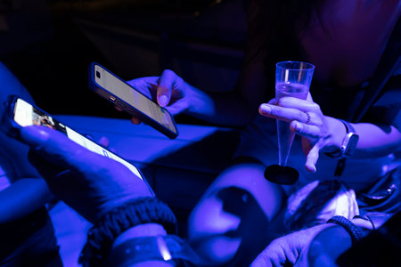 female hands holding a glass with an alcoholic drink and mobile phones, writing text, against the background and in the light of neon disco lighting of a nightclub.の写真素材