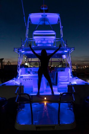 full length silhouette of a girl with raised arms and a cool gesture against the background of neon blue lights illuminating the territory of the catamaran yacht..の写真素材
