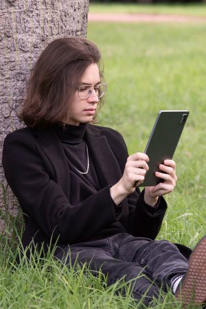 young student in business style clothes, wearing glasses, holding a micro tablet in his hands, working on the Internet online, sitting on the grass in a city park among the trees.Rest, work in the park.の写真素材