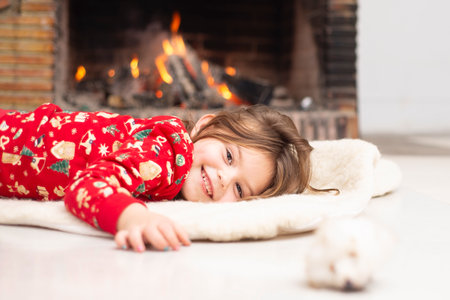 a little girl smiles sweetly, lies comfortably on a warm woolen carpet against the backdrop of a fire in the fireplace. Background For a project for the sale of warm winter accessories,for a fireplaceの写真素材