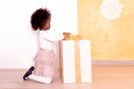 African Little girl plays with a bow color of the year 2024 peach fuzz on a large decorated gift box. Child opens a greeting box.Childrens partyの写真素材