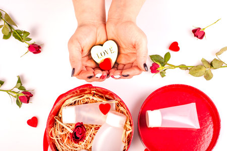 Women's hands hold a decorative heart next to a gift box with cosmetic products,with tubes of cream on the table with flowers and rose petals,hearts.Gift for a woman.Happy Women's Day. Cream in gift.の写真素材