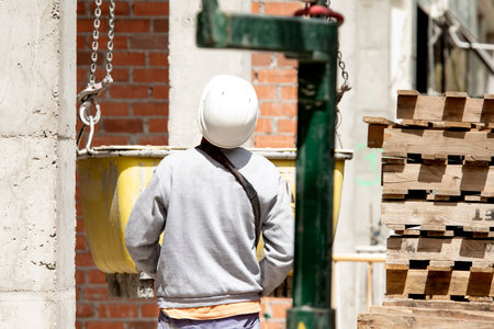 A builder in a hard hat watches the lifting of a container with a concrete mixture on a chain near a...