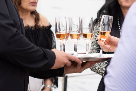 The waiter elegantly holds a tray with glasses of champagne with his hand, close-up, guests take wine glasses at a social event with people in the background. Service by party staffの写真素材