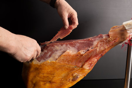 A man's hand cuts thin pieces of dried ham ham from a whole leg with a knife. Cooking Spanish food