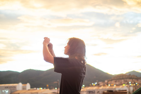 A person takes a picture of a picturesque sunset with mountains in the background on their smartphone during the golden hour. Harmony of technology and the environmentの写真素材