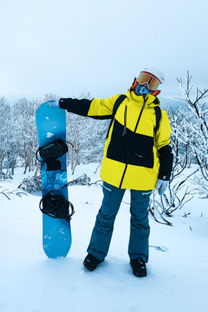A snowboarder in a colorful ski jacket and helmet holds a snowboard on a snowy slope with snowy mountains in the background. winter activity on holidayの写真素材