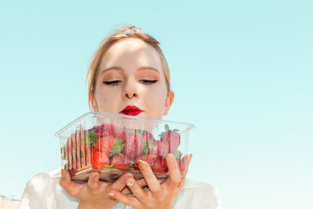Young woman holding a container with ripe strawberries. Enjoying fruits. Plastic disposable packaging with berries for food in human handsの写真素材