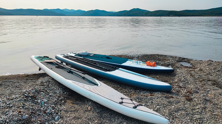 Three SUP boards with paddles on a rocky beach by the water with wooded hills and a quiet bay in the backgroundの写真素材
