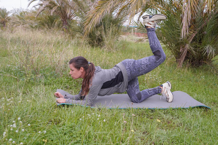 A woman doing gluteal exercises while balancing on one leg in a park lined with palm trees. Active recreation and sports. wellnessの写真素材