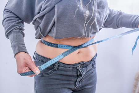 A person measures their waist with a measuring tape at home. Body size and body fat check. Weight loss and weight management conceptsの写真素材