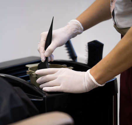 women stylist in gloves preparing color cream for dying hairin a container, a mask for treatment procedure in beauty salon. Hhairdresser mixing brush coloring hair concept supplies.の写真素材